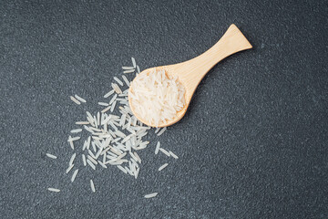 Wooden spoon filled with white rice and scattered grains on dark surface