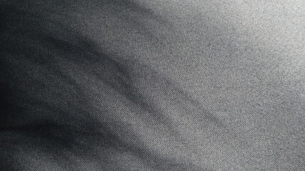 Photo of textured fabric of black color. From the upper left corner there is a shadow from the leaves of the plant. Thin long stripes. There is a light in the upper right corner. Monochrome.