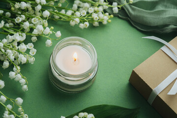 Glass candle burning with white lily of the valley flowers on green background
