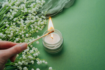 Lighting candle with match surrounded by white lily of the valley on green background