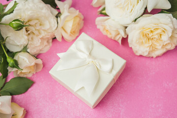 White gift box with ribbon surrounded by roses on pink background