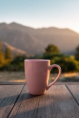 A blush-pink mug sits on a weathered wooden table, overlooking a sunlit mountain range at sunset