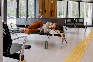 Tired young woman sleeping on airport, bus station bench with headphones and reusable bottle,...