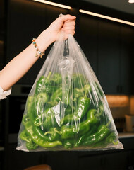 Womans Hand Holding a Transparent Bag Filled with Fresh Green Peppers Against a Dark Kitchen Background.
