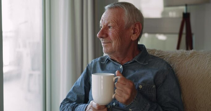 Peaceful positive retired senior man enjoying cup of morning coffee, holding mug of hot drink, sitting on comfortable couch, looking at window away, thinking, smiling, enjoying comfort
