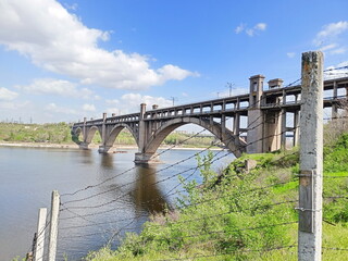 View from the forest thickets of Khortytsia Island on the Zaporizhzhia bridge across the Dnieper against the background of a sunny blue sky barely covered with snow-white clouds.