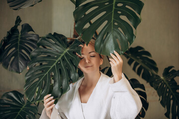 A young adult woman in the morning in a bathrobe with huge leaves of a monstera plant, emphasis on...
