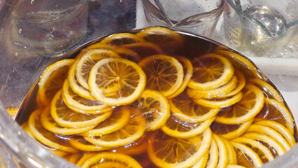 Thin lemon slices are submerged in a dark liquid, likely iced tea or syrup, inside a large clear...
