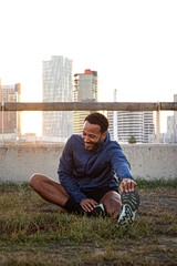 Smiling male runner stretching legs on rooftop with city view at sunset