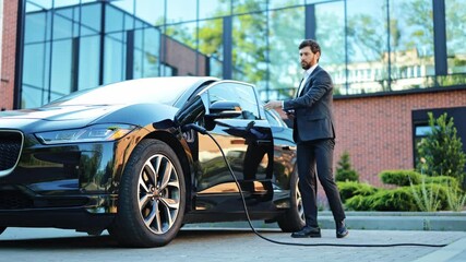 Professional businessman in a dark suit happily checks his smartphone while his sleek electric vehicle charges. Modern architecture provides the backdrop for this sustainable future.