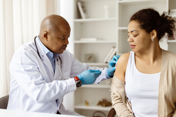 Female patient receiving injection from black doctor man wearing gloves during routine vaccination...