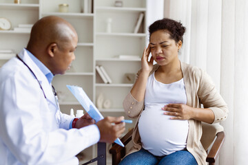Fototapeta premium Concerned pregnant woman discussing symptoms with her doctor during prenatal consultation. Expecting mother touching her head, possibly experiencing pain or discomfort
