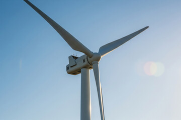 A close-up view of the nacelle and blades of a modern wind turbine against a clear blue sky, with the sun slightly to the right.