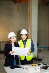 Construction engineer in a safety vest and an architect with a hard hat examine blueprints and a scale model on a table, collaborating