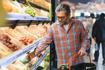 Stylish senior man in red shirt choosing vegetables and fruits at supermarket