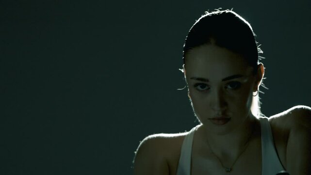 Kobieta trenująca na rowerze stacjonarnym, widziana od frontu. Światło z tyłu podkreśla jej ramiona, włosy i głowę. Porusza się z gracją i zalotnie patrzy w stronę kamery, emanując pewnością siebie