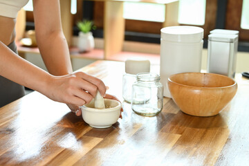 A woman is preparing a mixture in a bowl on a wooden table. The table is cluttered with jars, a bowl, and a bottle. The woman is focused on her task, and the scene conveys a sense of concentration