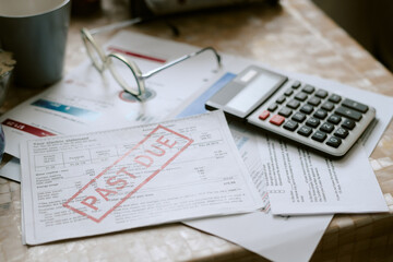 Senior Caucasian couple struggling with overdue utility bills, holding documents marked past due on cluttered table with calculator and eyeglasses, financial hardship evident