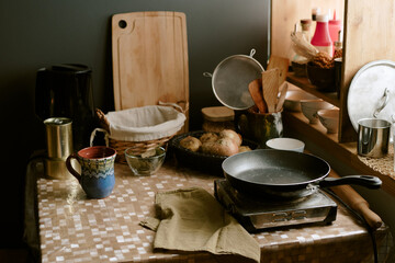 Kitchen table displaying frying pan on portable stove, ceramic mug, bread rolls in basket, wooden utensils, cutting board, metal kettle, onions, and various kitchenware
