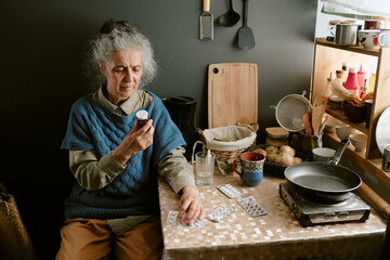 Senior Caucasian woman sitting at kitchen table holding medication cup, examining pills with...