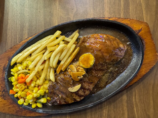 A sizzling plate of steak with gravy, french fries, corn, carrots, and mushrooms on a wooden table.