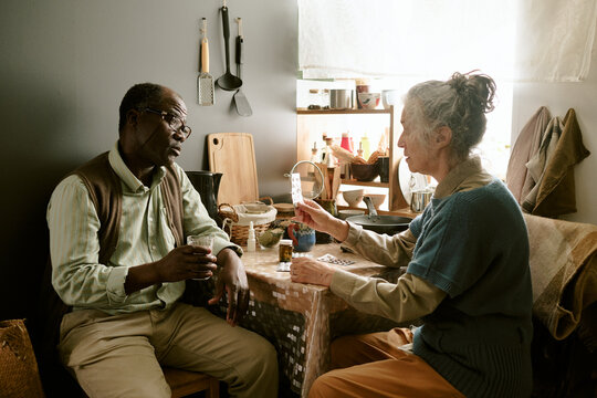 Senior Caucasian woman handing medication to senior Black man while sitting together at small kitchen table, both appearing engaged in conversation, daily life scene of elderly couple