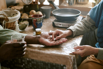 Senior Black man receiving medication from senior Caucasian woman at kitchen table, hands visible...
