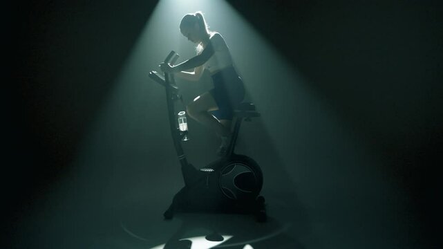 Kobieta wsiadająca na rower stacjonarny w czaciemnionej sali z oswietleniem filmowym 