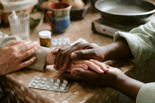Senior Caucasian woman and senior Black man sitting at table holding hands, showing support and care, with medication blister packs and prescription bottle visible on tabletop
