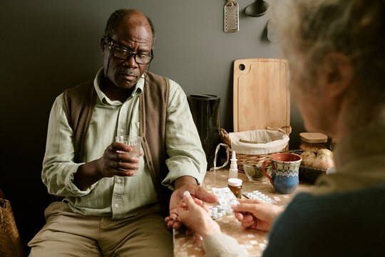 Senior Black man sitting at table holding glass of water while senior Caucasian woman handing medication, both engaged in daily routine, kitchen background visible, poverty context evident - Powered by Adobe