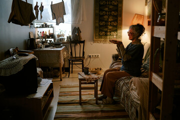 Senior Caucasian woman sitting on sofa holding hands together in small modest living room, watching...