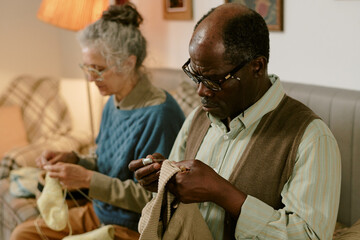Senior Caucasian woman and senior Black man sitting together knitting, focusing on handmade...