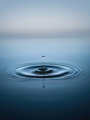 A single water droplet hangs suspended above a calm, still surface, creating concentric ripples that expand outward. The image's muted blue tones evoke serenity