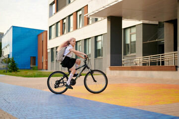 young girl riding bicycle in urban area, wearing school uniform and backpack. vibrant colors of...