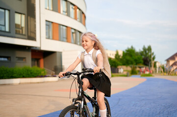 young girl riding bicycle near school building, smiling with joy on sunny day. wearing backpack,...