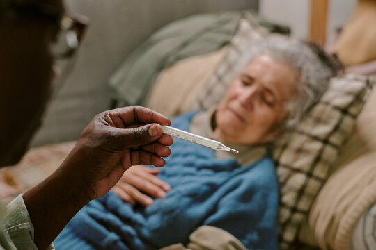 Senior Caucasian woman lying in bed with eyes closed, senior Black man holding thermometer in foreground, both appearing concerned, illustrating elderly couple facing health challenges