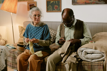 Senior Caucasian woman knitting beside senior Black man mending clothing while sitting on couch,...