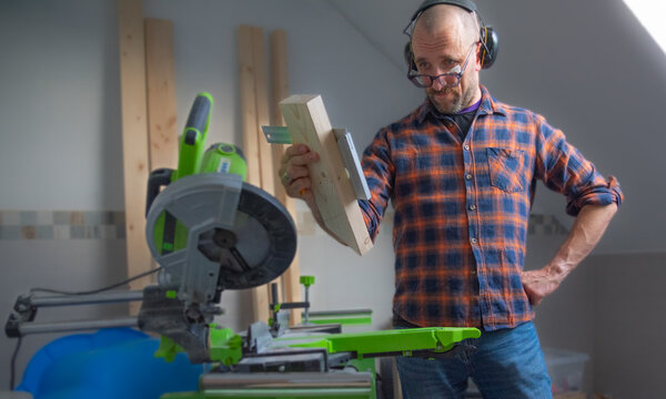 A carpenter, white man with protective eyeglasses and ear protection standing next to a miter saw and measuring the next cut with a metal angle