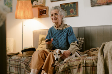 Senior Caucasian woman sitting on sofa holding remote control and petting small dog, wearing...