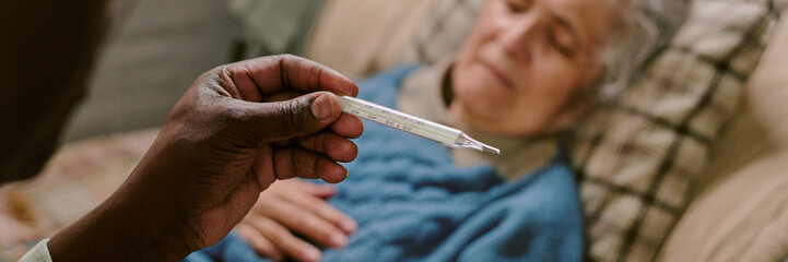 Senior Caucasian woman lying on couch with eyes closed, Black man holding thermometer in foreground...