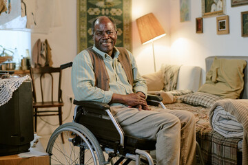 Senior Black man sitting in wheelchair smiling in modest living room, hands resting on lap,...