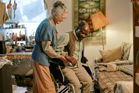 Senior Caucasian woman assisting senior Black man with disability transferring from wheelchair to bed in modest home setting, both focused on movement, demonstrating care and support