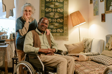 Senior Caucasian woman standing behind senior Black man sitting in wheelchair, both smiling, woman resting hand on mans shoulder, couple posing together in modest home interior
