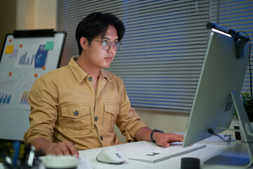 Focused man in glasses working late on desktop computer in a dimly lit office