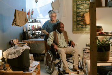 Senior Caucasian woman standing behind senior Black man sitting in wheelchair with disability, both...