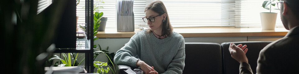 Woman sitting on sofa during therapy session, looking down with thoughtful expression while...