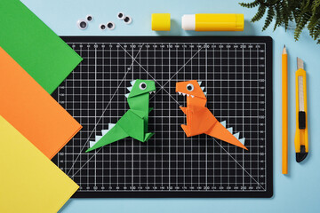 Green and orange origami dinosaurs on a black cutting mat. Surrounded by colorful sheets of paper,...