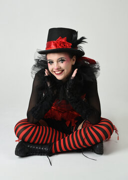 Burlesque circus performer, fantasy female model in halloween jester clown costume with top hat, red gothic corset. theatrical sitting pose kneeling on floor, isolated white studio background.