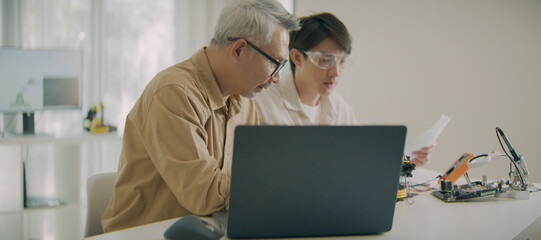 Senior and young Asian engineers collaborating on electronics project with laptop and circuit...
