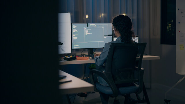 A dedicated female programmer seen from behind, intensely coding across multiple monitors in a dark office. Focuses on AI, machine learning, and software development.